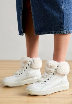 Hvite skinnhøye sneakers med plysj-fôr på mansjettene, sølvfargede hull og en glidelås på siden, satt mot et tregulv.