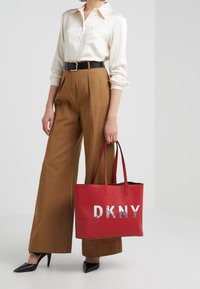 Röd syntetisk tote-väska med silverfärgad "DKNY"-logotyp, slät textur, långa handtag, uppställd med beige högmidjade byxor och en krämfärgad skjorta.