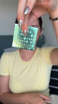 Mano con unghie argento metallizzato che tiene un illuminante compatto verde MAC Bounce & Gleam davanti a una persona che indossa una maglietta gialla e orecchini a cerchio.