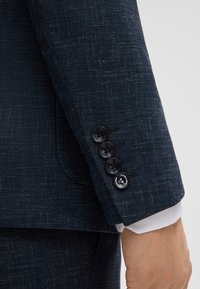 Maniche di una giacca da abito blu navy con una finitura testurizzata, caratterizzata da quattro bottoni neri e una manica di camicia bianca visibile sotto.