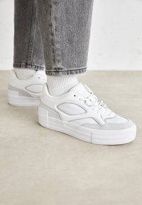 Vit sneaker med slät läderovandel och grå mockadetaljer, en smidig, rundad design och en tjock, vit gummisula.