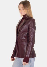 Blazer in pelle bordeaux con design aderente, revers a incavo, tasche frontali e polsini in maglia a coste. Texture liscia con leggero splendore.
