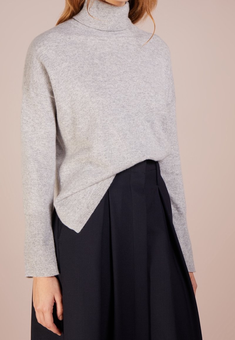 Pull à col roulé gris avec une coupe décontractée. Texture tricotée, manches longues et fentes latérales. Associé à une jupe noire, créant un look superposé.