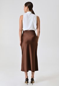Gonna midi in satin marrone con una texture liscia, abbinata a una blusa bianca senza maniche. Vita aderente e silhouette svasata, indossata con tacchi neri.