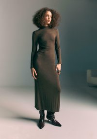 Rjava midi obleka z dolgimi rokavi in prilegajočim krojem, narejena iz mehkega materiala. V kombinaciji z oštrimi črnimi usnjenimi škornji za kontrast.