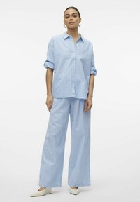 Camicia button-up a strisce azzurro chiaro e bianca con maniche arrotolate, abbinata a pantaloni a gamba larga abbinati, caratterizzati da una vestibilità rilassata e una texture morbida.