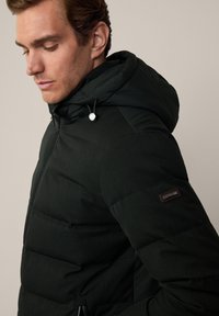 Giacca imbottita in tessuto verde scuro con cappuccio. Presenta una chiusura con zip, una superficie testurizzata e una piccola etichetta con marchio sulla manica.