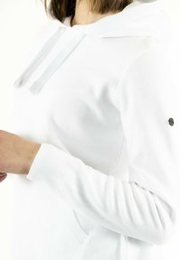 Persona che indossa un felpa bianca con cappuccio, maniche lunghe, polsini a coste e un piccolo logo rotondo sulla parte superiore del braccio.