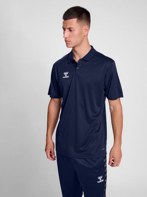 Joven con el pelo corto que lleva una camiseta polo azul marino y pantalones a juego con logos de hummel, de pie y mirando a su izquierda.