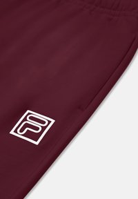 Burgundowe spodnie sportowe z elastycznym pasem i białą łatą z logo. Gładki materiał bez widocznych wzorów czy detali hardware'owych.