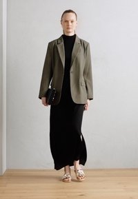 Blazer vert olive sur une robe longue noire avec une fente sur le côté, assorti à des sandales à lanières de couleur claire et tenant une pochette noire. Fond uni.
