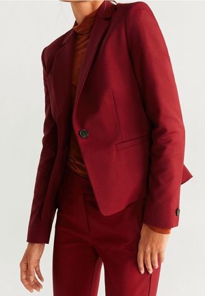 Kvinde iført en tætsiddende mørkerød blazer og matchende højtaljede bukser over en rustfarvet rullekravebluse.