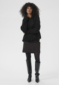 Kvinde med krøllet hår iført en sort tyk sweater, mørkebrun nederdel, sorte strømpebukser og knæhøje sorte støvler, som står med den ene hånd nær sit ansigt.