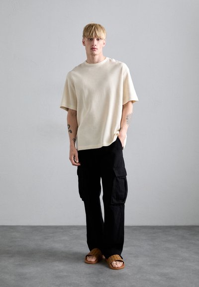 Chemise à manches courtes en tricot côtelé crème, coupe décontractée ; associée à un pantalon cargo noir avec de grandes poches ; sandales à glissière brunes avec perforations.