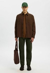 Ung mand iført en grøn hat, brun overskjorte, brun sweater, grønne bukser, sorte sko, holder en brun tote bag, stående på en ensartet hvid baggrund.