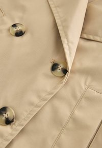 Beige trenchcoat stof met een gladde textuur, voorzien van grote, donkere schildpadknopen en een gelamineerde kraag.