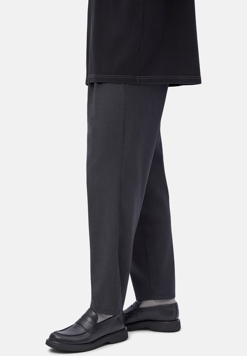Hombre vestido con pantalones oscuros de corte ajustado y mocasines de cuero negro, de pie frente a un fondo liso y claro.