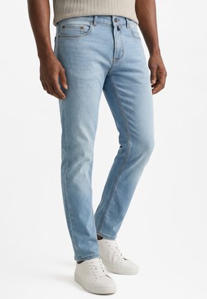 Mann trägt hellblaue, enganliegende Jeans und weiße Sneaker und steht vor einem schlichten weißen Hintergrund.