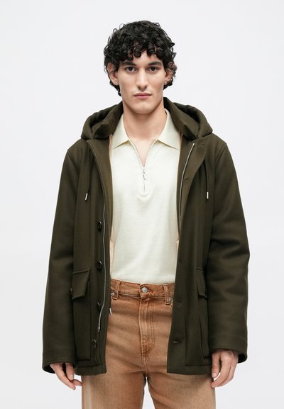 Oliivinvihreä hupullinen takki, jossa napit ja vetoketjut, yhdistetty vaaleaan vetoketjulliseen poolopaitaan ja haalistuneisiin ruskeisiin housuihin.