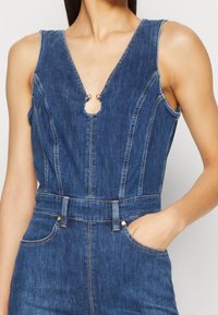 Mono de mezclilla con escote en V profundo y detalle de corte, que presenta costuras en contraste, una cintura ajustada y bolsillos laterales.
