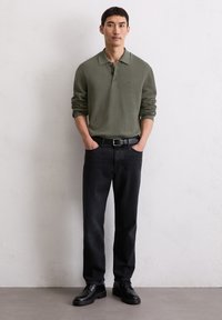 Polo shirt a maniche lunghe verde oliva con una finitura texturizzata, pantaloni in denim neri, cintura nera e scarpe stringate nere. In piedi contro uno sfondo neutro.