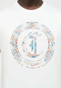 Κοντινή λήψη λευκού T-shirt με πολύχρωμο λουλουδάτο κυκλικό λογότυπο και το κείμενο «Just Cavalli» στο στήθος.
