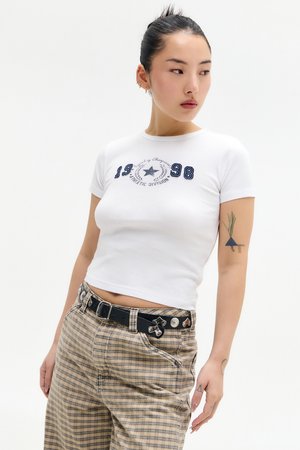 Camiseta blanca ajustada con bordado en azul marino que muestra "1998" y una estrella, combinada con pantalones de cuadros beige y un cinturón negro con detalles metálicos.