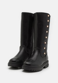 Bottes longues noires en cuir lisse, avec un bout rond, une semelle texturée et des clous décoratifs en forme de cœur en or rose le long du côté.