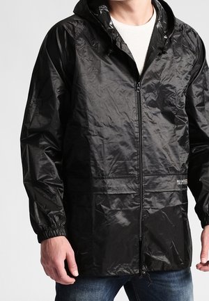 Leichte schwarze Regenjacke mit Kapuze, Frontreißverschluss und elastischen Bündchen, getragen über einem weißen Hemd und blauen Jeans.