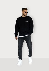 Schwarzer Strickpullover mit Rundhalsausschnitt und geripptem Saum, über einem weißen Hemd getragen, kombiniert mit dunklen Jeans und weißen Sneakers. Runde Sonnenbrille inklusive.