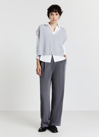 Gilet sans manches en tricot gris clair avec un décolleté en V et des boutons sur le devant, superposé sur une chemise blanche à col et associé à un pantalon large gris.