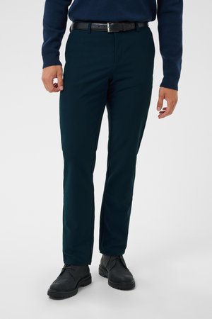 Hombre con pantalones azul marino oscuro, cinturón negro, botas negras y un suéter azul marino de manga larga de pie sobre un fondo claro y liso.