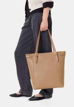 Persona con jeans grises y zapatos planos negros sosteniendo un bolso tote de cuero beige con herrajes dorados al costado.