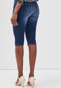 Shorts en denim bleu foncé, arrivant au genou, avec une coupe ajustée et une légère élasticité. Comprend des poches arrière et des détails couture à l'ourlet.