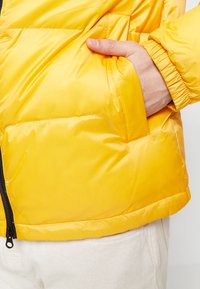 Gelbe Pufferjacke mit glatter, glänzender Textur. Verfügt über eine Seitentasche und einen schwarzen Reißverschluss am Saum für zusätzliche Funktionalität.