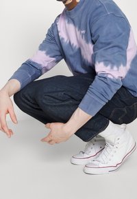 Felpa crew tie-dye azzurro chiaro e viola, jeans in denim scuro e sneakers alte bianche con dettagli rossi. Look semplice e casual.