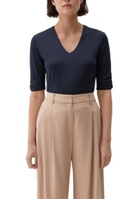 Marineblaue V-Ausschnitt-Bluse mit Dreiviertelärmeln, glatter Textur, kombiniert mit hochtaillierten, beigen, weit geschnittenen Hosen mit Falten.