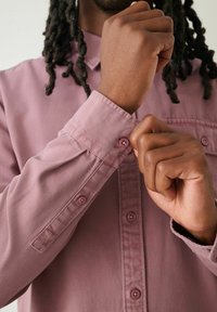 Langärmliges, staubrosa Hemd aus einem weichen Stoff mit Knopfmanschette und Kragen. Nahaufnahme von Händen, die den Knopf am Handgelenk des Hemdes anpassen.