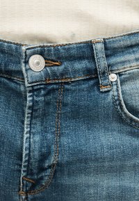 Κοντινό πλάνο της μέσης ενός μπλε τζιν τζιν, που δείχνει μεταλλικό κουμπί, θηλιά ζώνης, άκρη μπροστινής τσέπης και πορτοκαλί ραφές λεπτομέρειες.