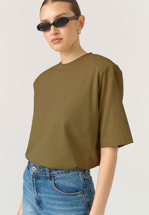 Olívazöld, oversized t-póló, puha anyagból, rövid ujjakkal és kerek nyakkivágással, világoskék farmer rövidnadrággal párosítva.
