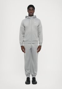 Lysegrå træningsdragt lavet af blødt stof. Den lynlåsede hoodie har snoretræk og logo. Bukserne har en afslappet pasform og har elastiske manchetter samt lommer.