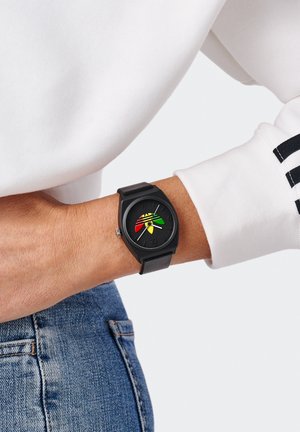 Fekete számlapos karóra fekete szíjon, piros, sárga és zöld absztrakt mintával, fehér hosszú ujjú felsővel és kék farmerral viselve.