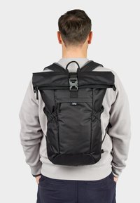 Zaino nero con design a roll-top, spallacci regolabili, chiusura con fibbia e tasche laterali; realizzato in tessuto resistente con una superficie liscia.