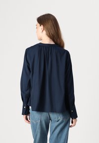 Camicia a maniche lunghe blu navy realizzata in tessuto morbido, caratterizzata da un collo arricciato, polsini con bottoni e una vestibilità rilassata e svasata.