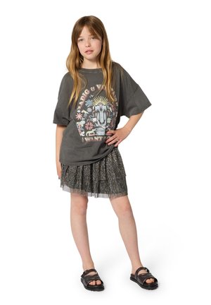 Szara oversize koszulka z nadrukiem graficznym, warstwowa metaliczna plisowana spódnica oraz czarne sandały z regulowanymi paskami.