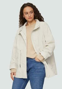 Lichtbeige oversized jasje met een rits en drukknoopsluiting, voorzien van twee klepzakken en een koord in de taille. Gemaakt in combinatie met een beige trui en jeans.