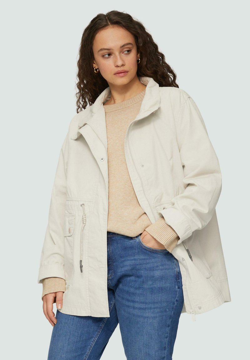 Lichtbeige oversized jasje met een rits en drukknoopsluiting, voorzien van twee klepzakken en een koord in de taille. Gemaakt in combinatie met een beige trui en jeans.