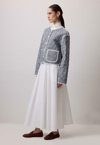 Prešívaná sivá bunda s bielym lemovaním, s vysokým golierom, zapínaním na gombíky a predným vreckom, nosená cez bielu, priedušnú sukňu.