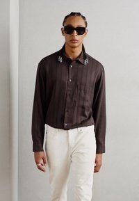 Chemise à manches longues marron à rayures avec une poche, col orné, associée à un jean blanc. Modèle portant des lunettes de soleil noires.