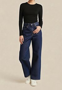 Haut noir à manches longues associé à un jean denim bleu foncé taille haute à jambes larges. Le jean présente un style classique à cinq poches et une fermeture par bouton.